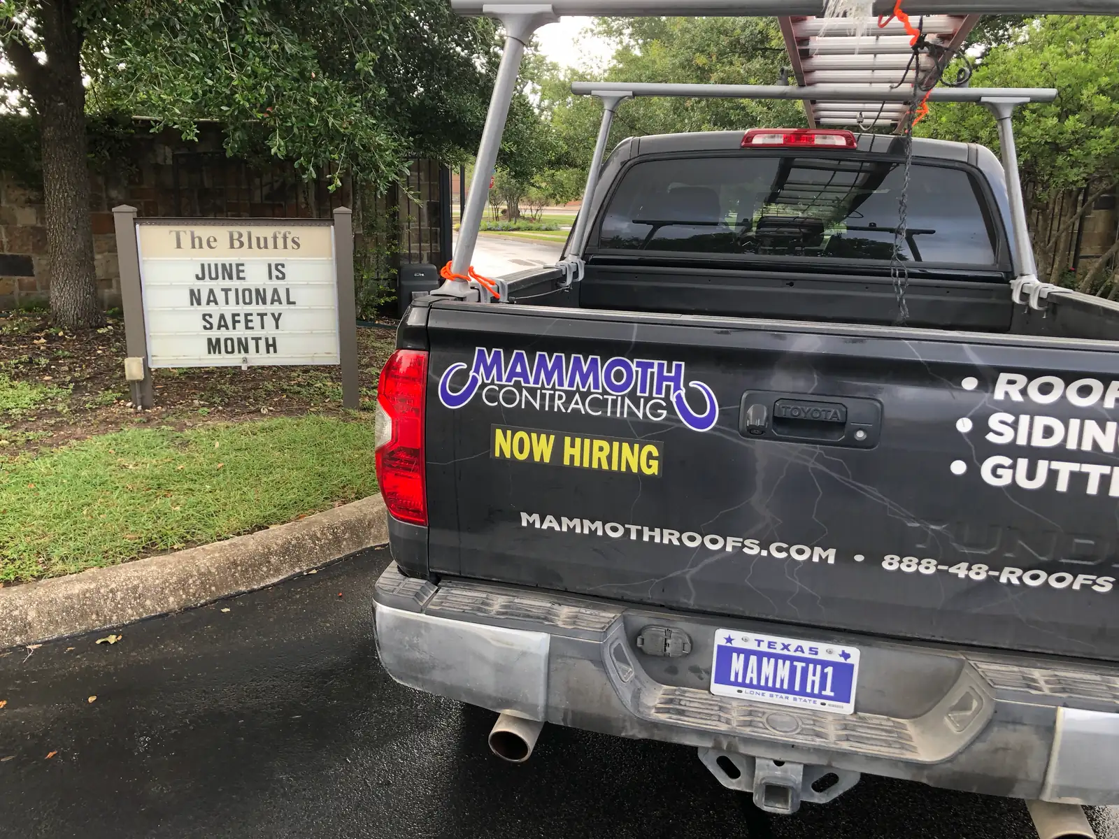 The tailgate of a wrapped Mammoth Contracting truck reading Now Hiring, parked beside a church's roadside sign