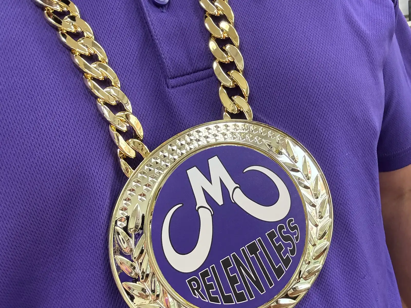A gold medallion on a chain, engraved with the Mammoth M and the word Relentless, worn over a purple Mammoth shirt