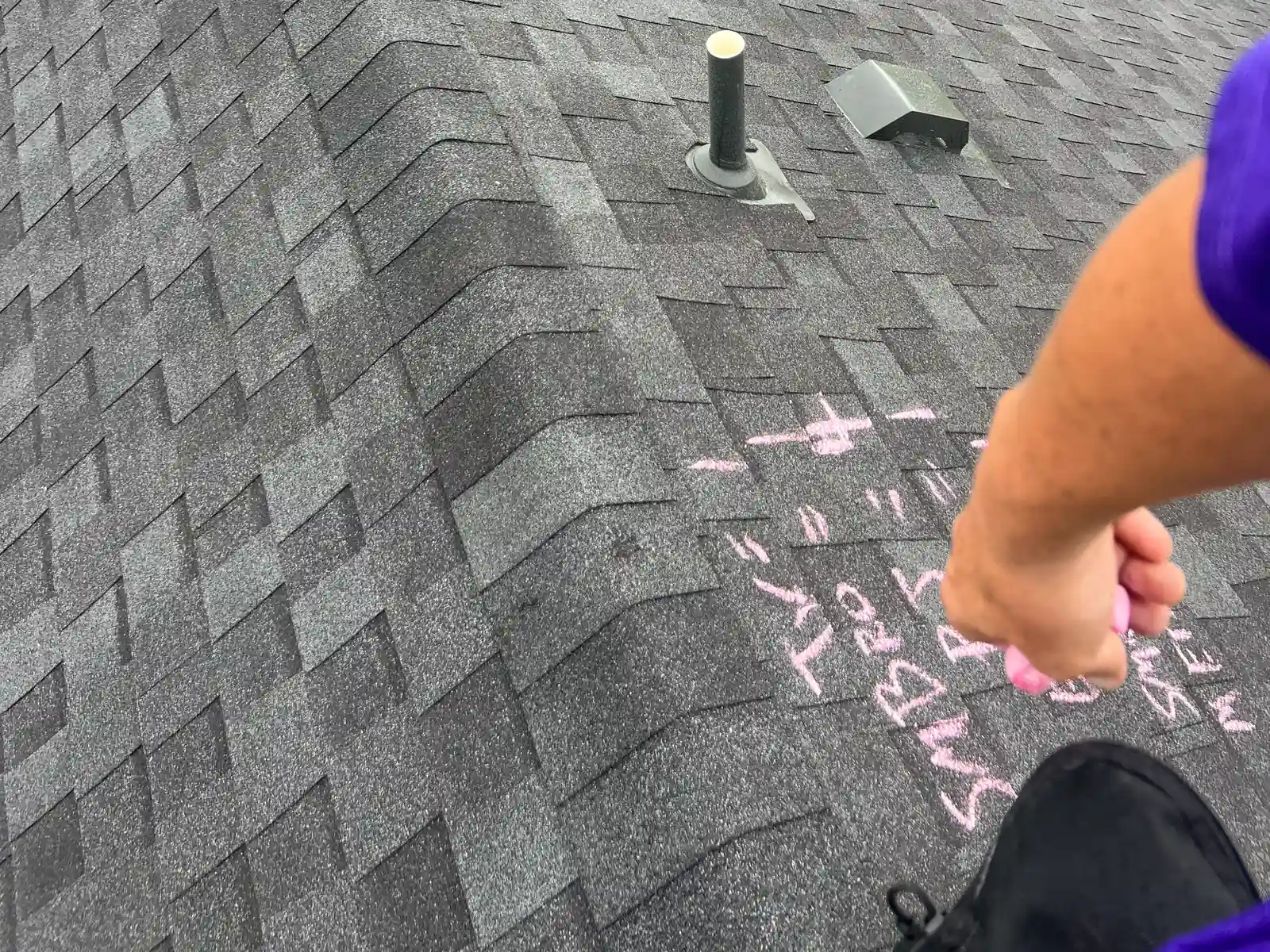 An inspector in a purple shirt writes a damage tally in pink chalk on a shingle roof slope near two roof vents
