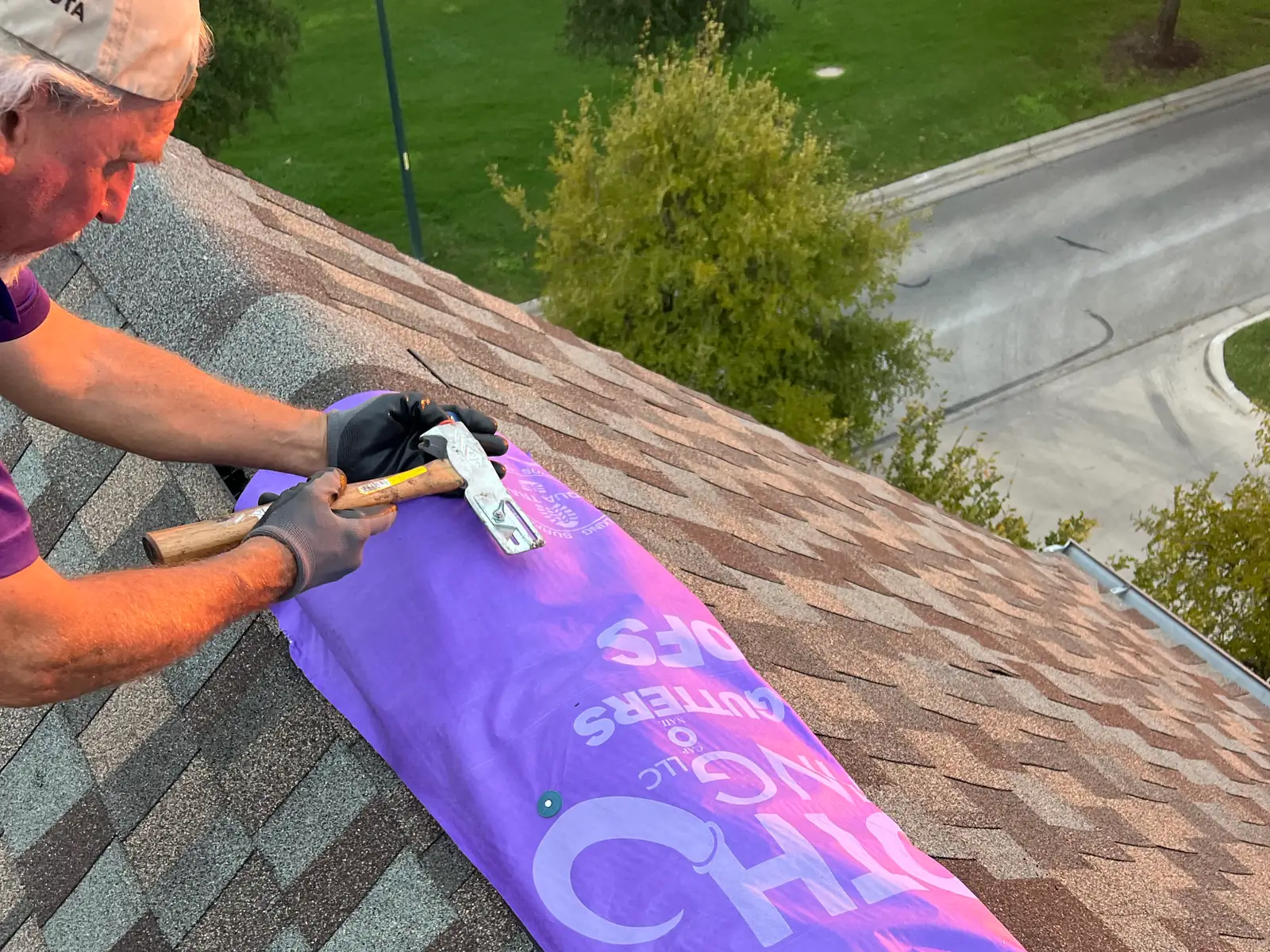 Gloved hands hammer cap nails through a purple Mammoth tarp along a roof slope
